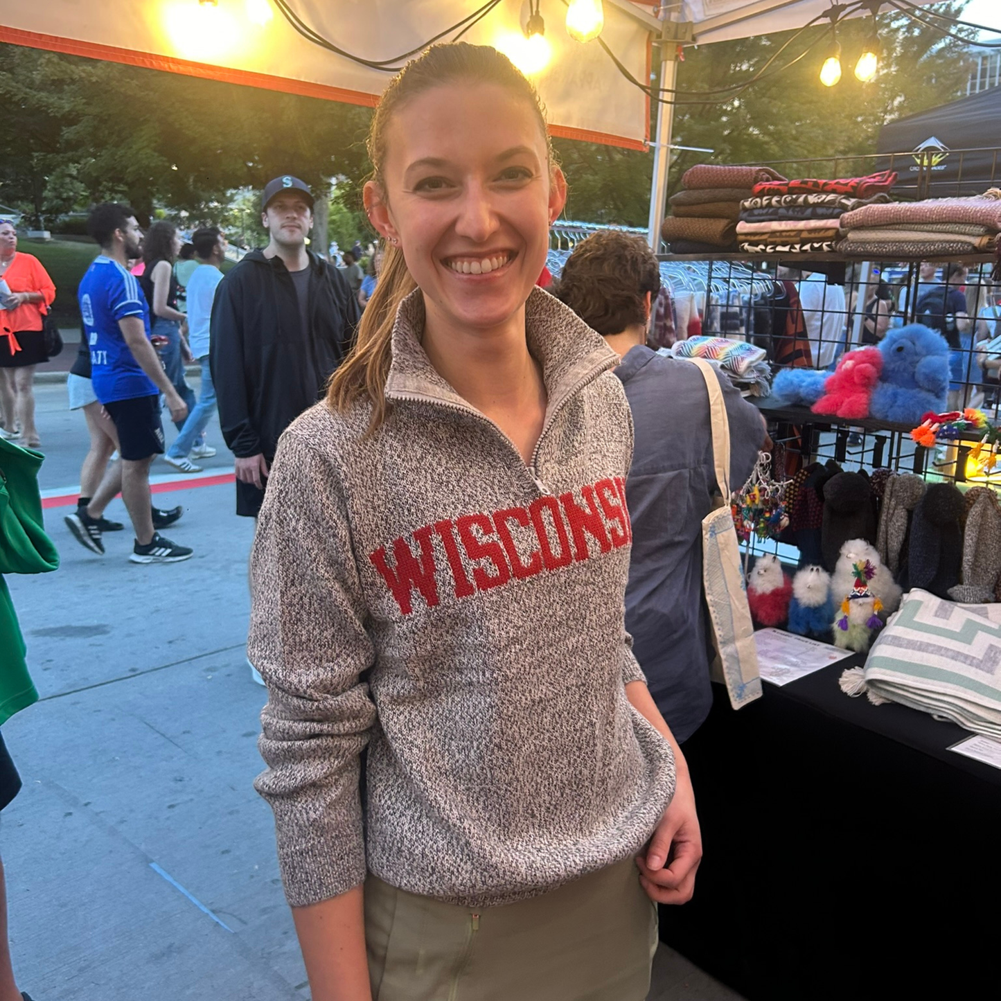 Woman wearing a 'WISCONSIN' sweater standing outdoors with people and booths in the background.
