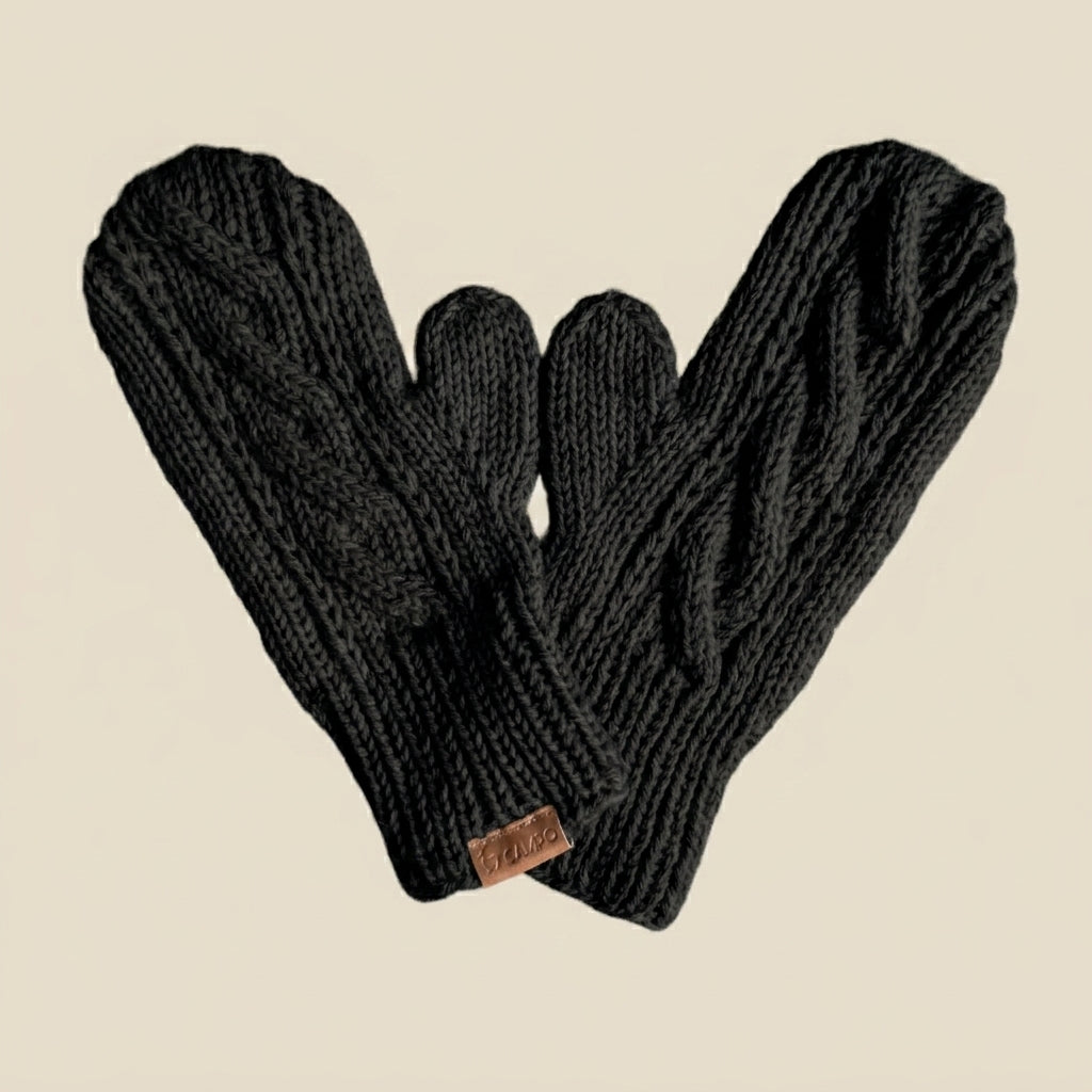 A pair of black cable-knit fingerless mittens arranged in a heart shape against a solid beige background. A small brown leather brand tag is visible on the cuff.