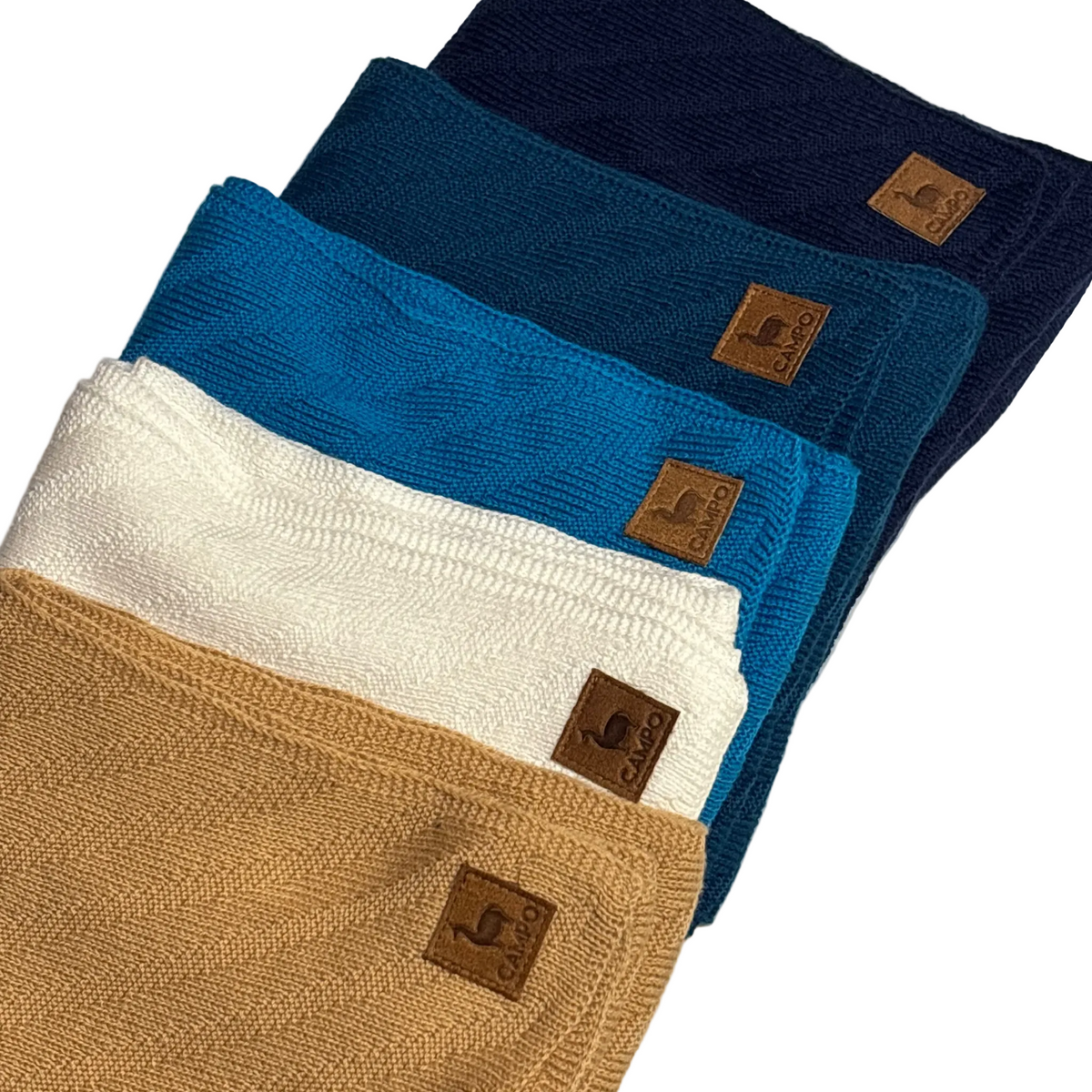 Four textured woven cloth napkins in navy, teal, cream, and tan, arranged diagonally on a light gray surface. Each napkin has a small brown leather logo patch with the brand name 'COCO'.