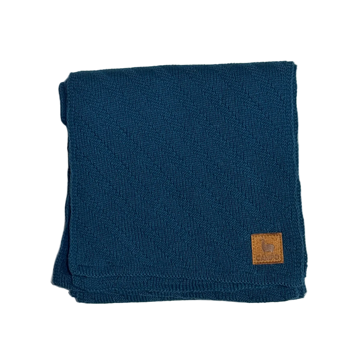 A folded dark teal knit blanket with a small tan leather brand patch on the bottom right corner, set against a plain white background.