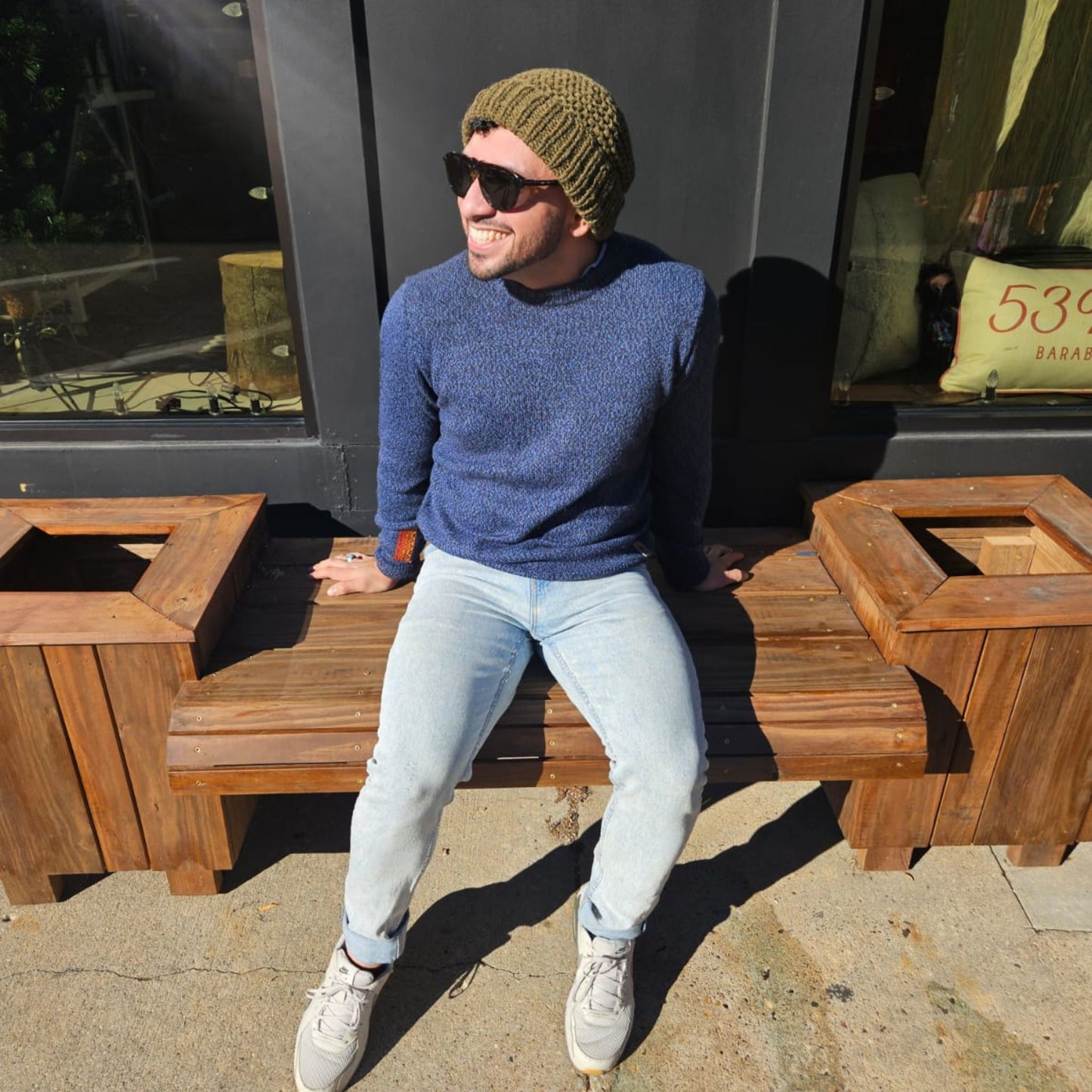 Man sitting on a wooden bench wearing a blue sweater, light jeans, and a green beanie.