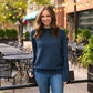 Woman wearing a blue sweater and jeans standing outdoors with tables and chairs in the background.