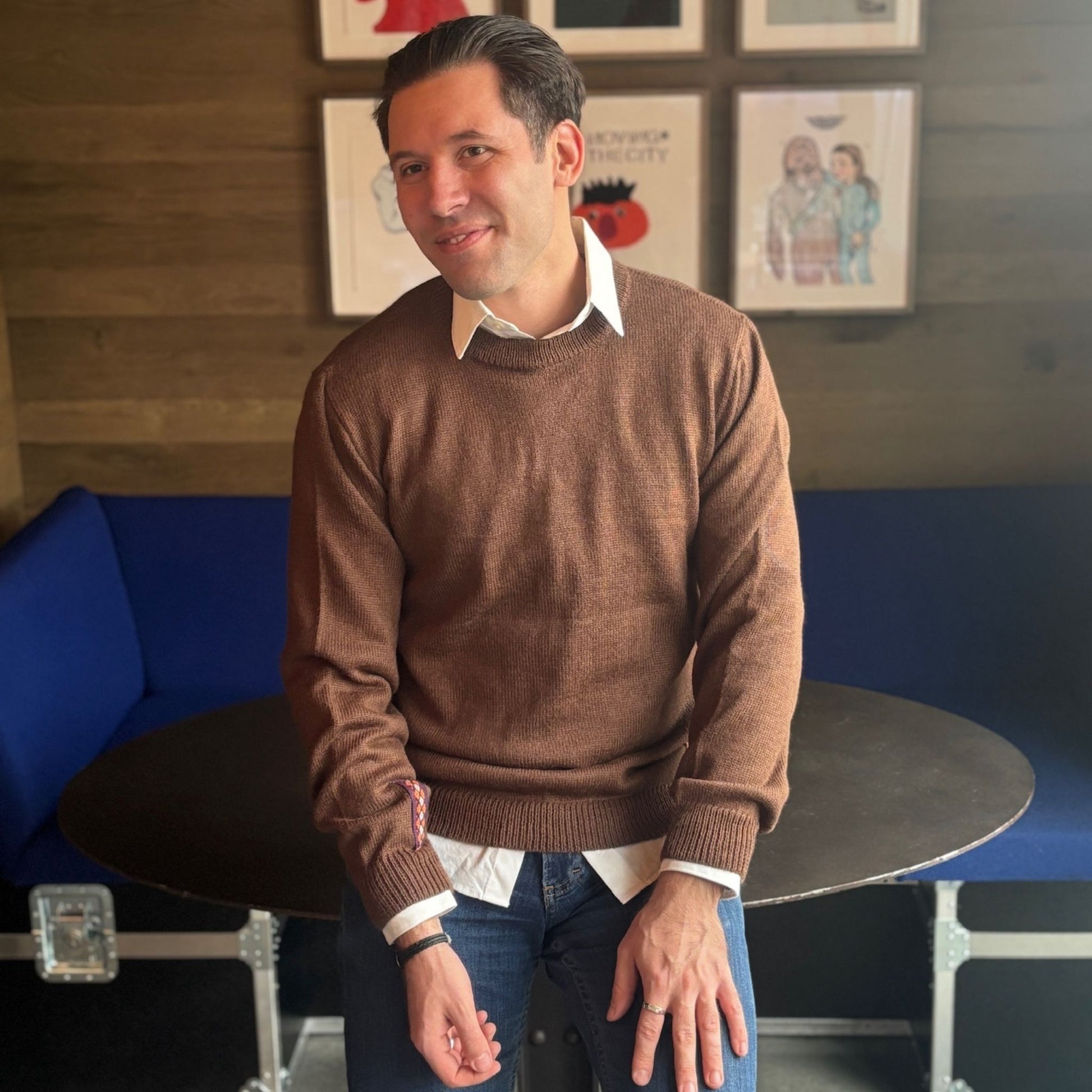 Man wearing a brown sweater in a room with blue couch and framed pictures on the wall.