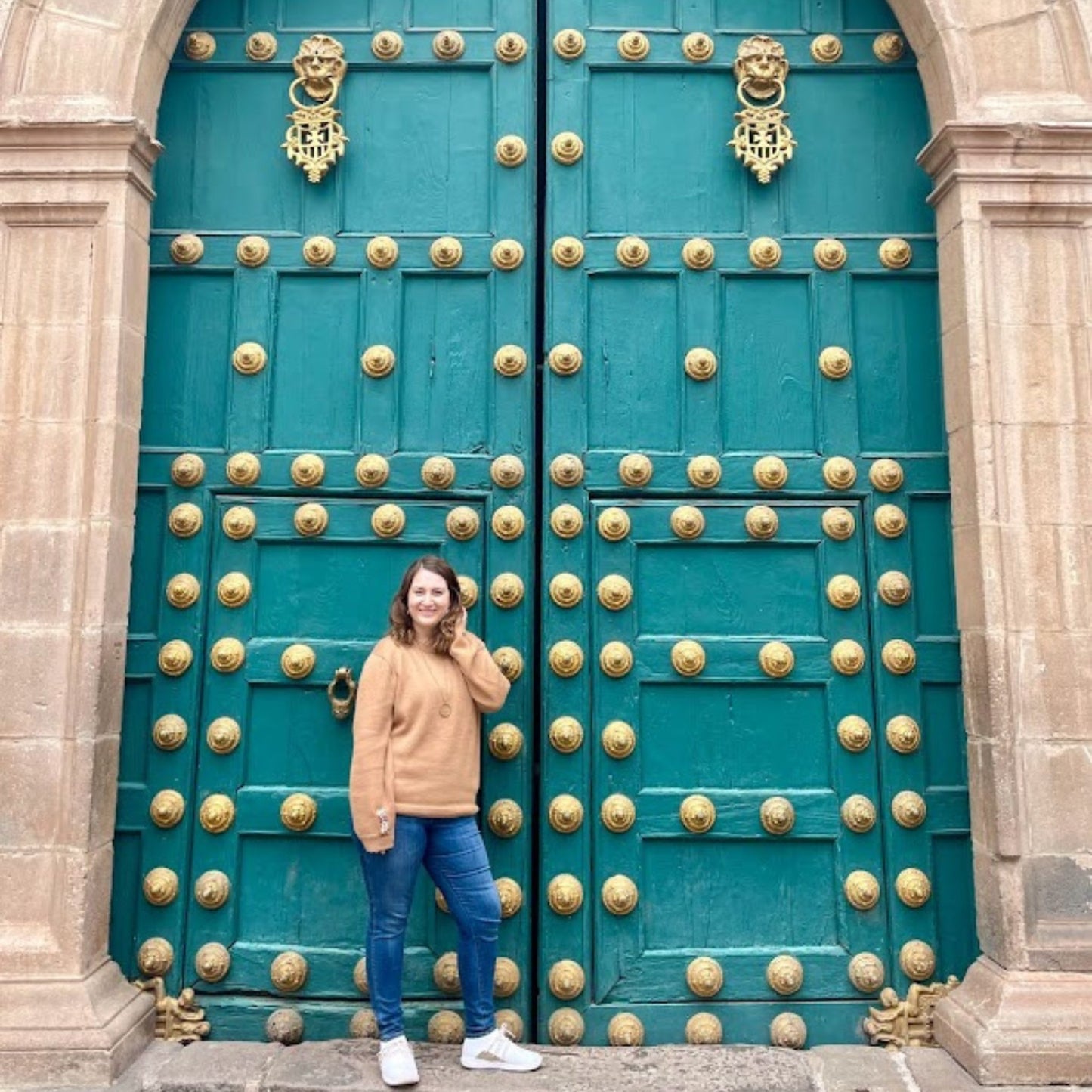 Woman wearing hazelnut  standing in front of a large teal door with gold embellishments