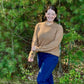 Woman in a hazelnut sweater and blue pants standing in a forest