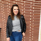 Woman wearing a black leather jacket, salt and peppre sweater, and blue jeans standing in front of a brick wall.