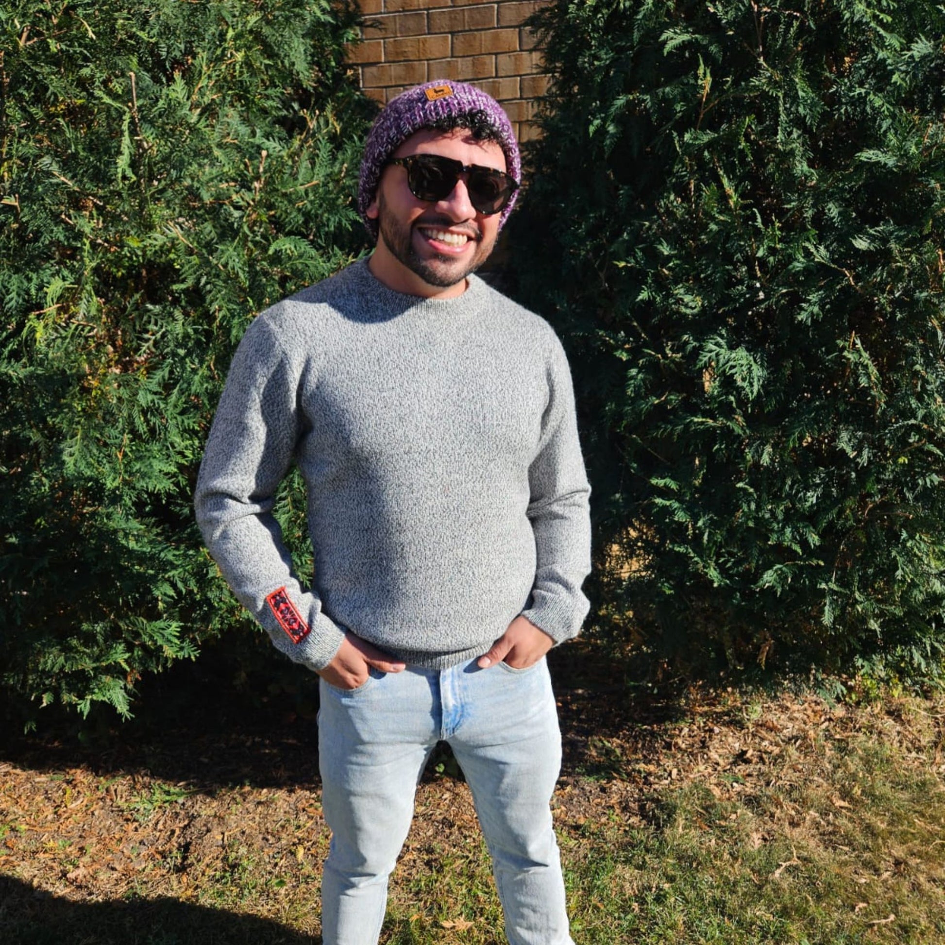 Man wearing a salt and pepper sweater, light blue jeans, and a purple beanie standing outdoors with greenery in the background.