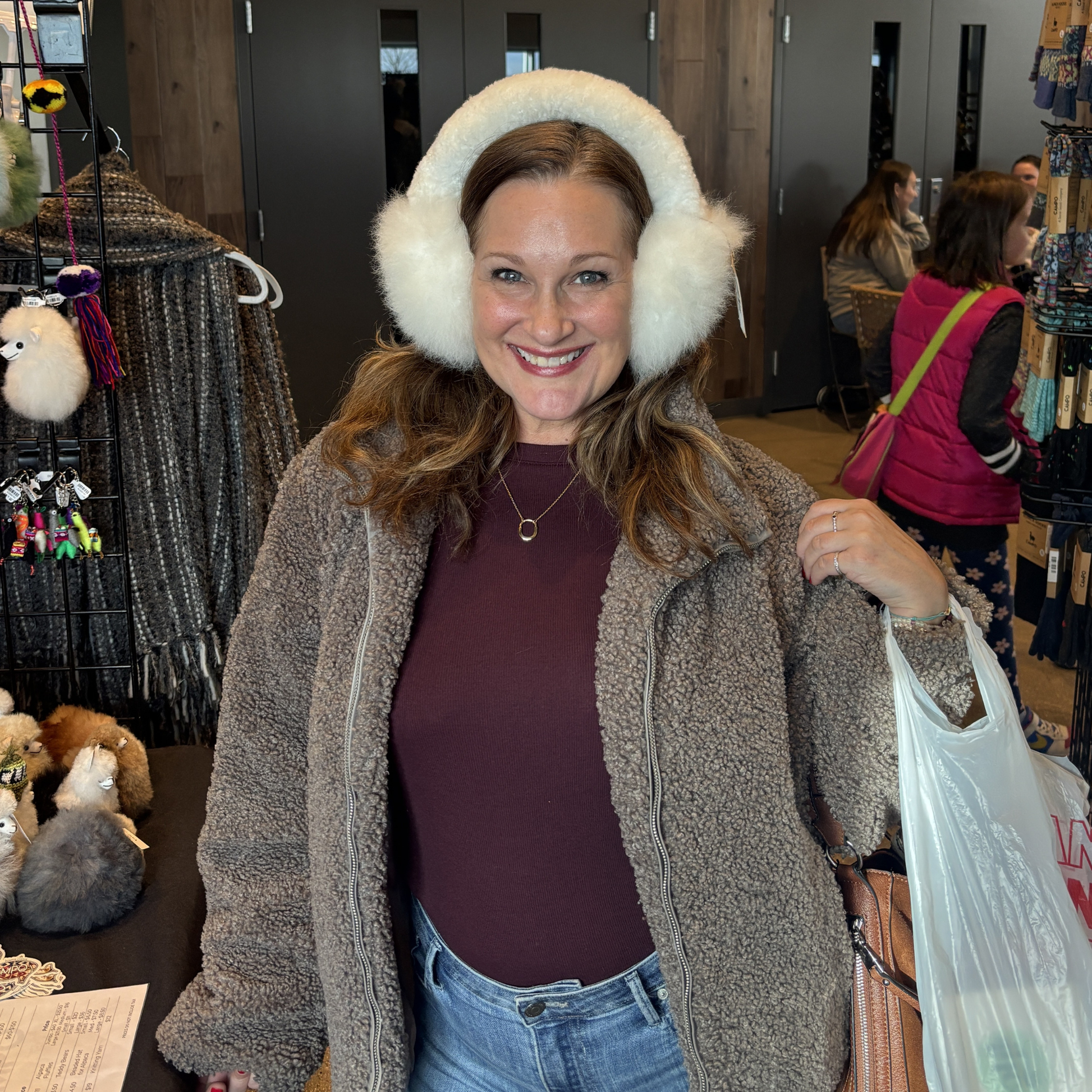 Woman wearing white fur ear muffs and a brown jacket in a store setting.