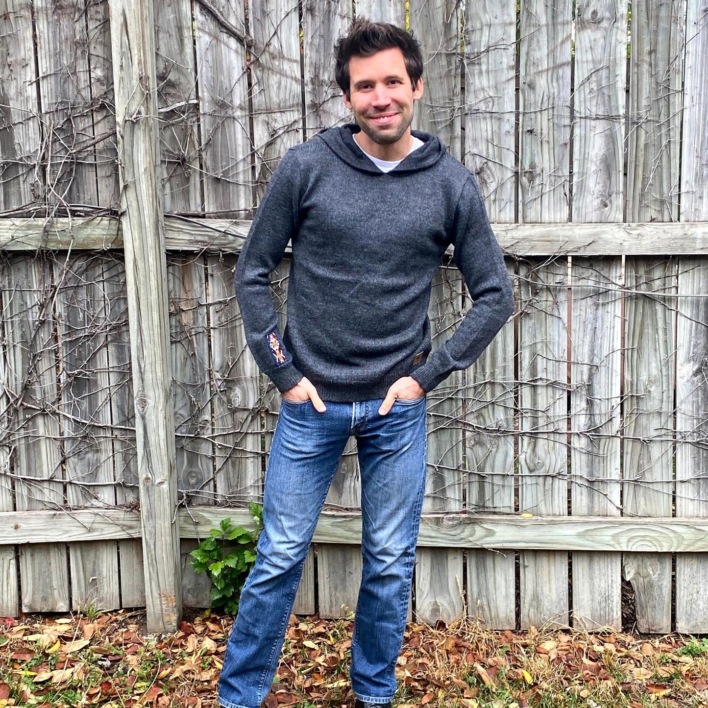 Man wearing a dark gray sweater and blue jeans standing against a wooden fence.