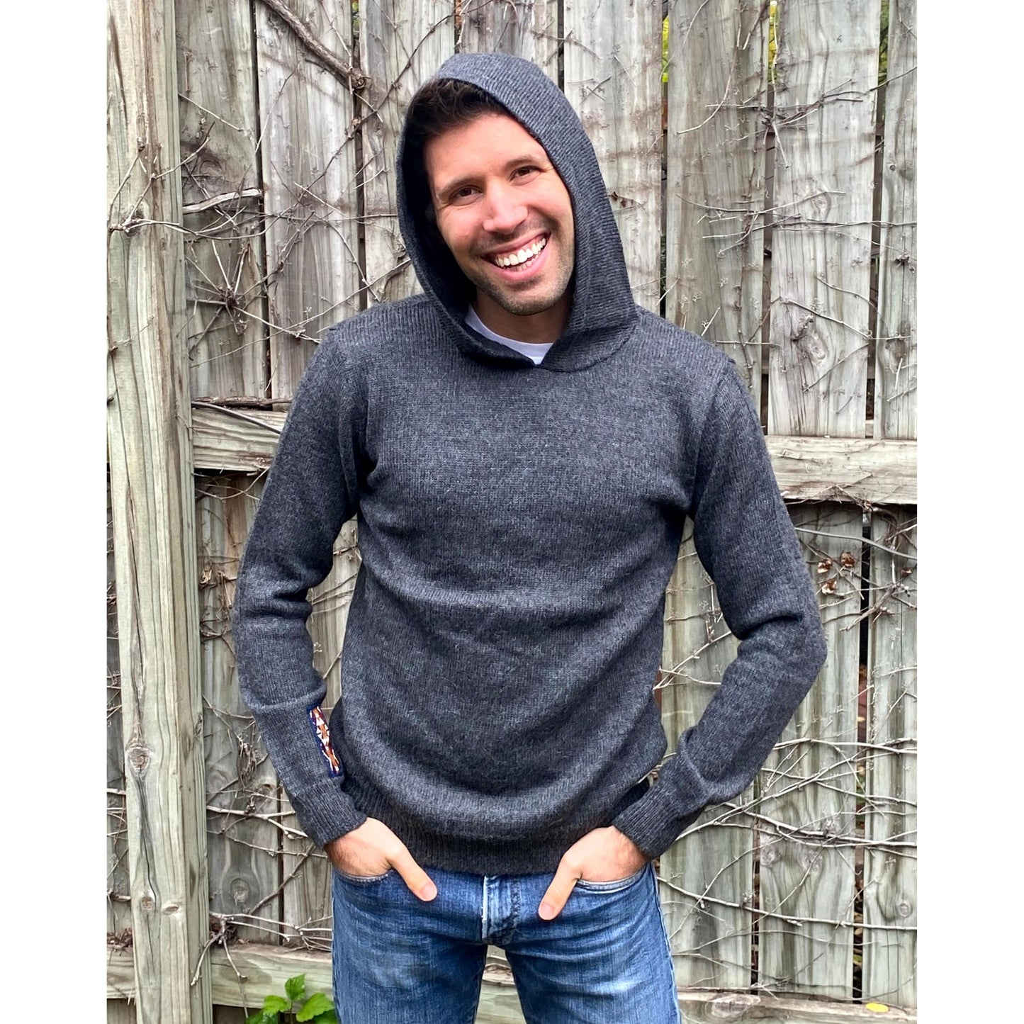 Man wearing a dark gray hoodie and jeans standing in front of a wooden fence.