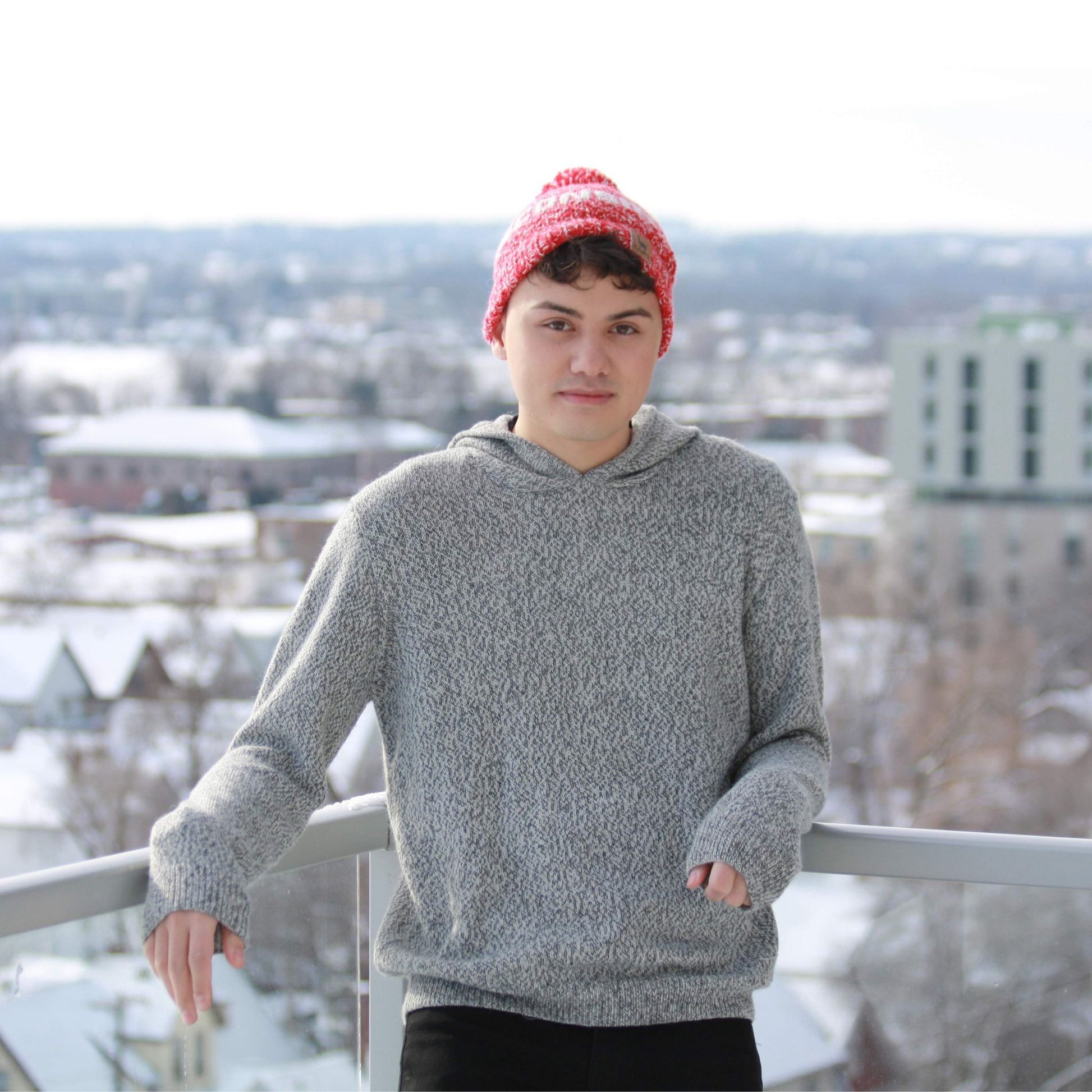 Person wearing a salt and pepper hoodie and red beanie standing on a balcony with a snowy cityscape background