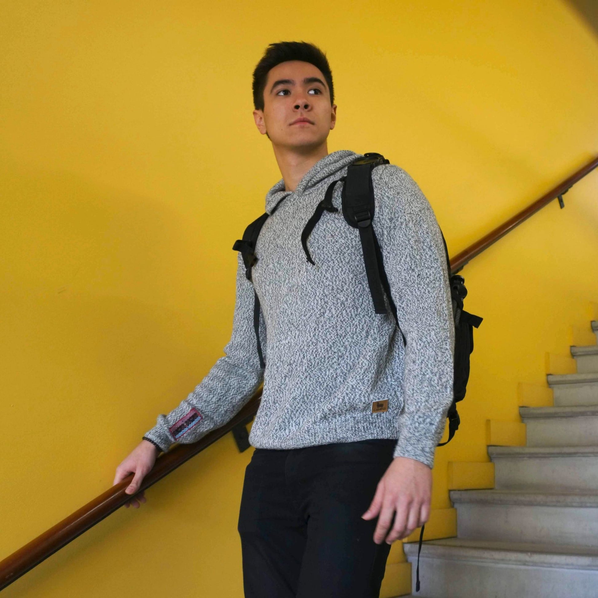 Person wearing a gray hoodie and black backpack against a yellow wall.