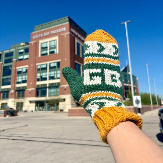 Knitted glove with 'GB' design held up in front of a building with 'Green Bay Packers' branding.