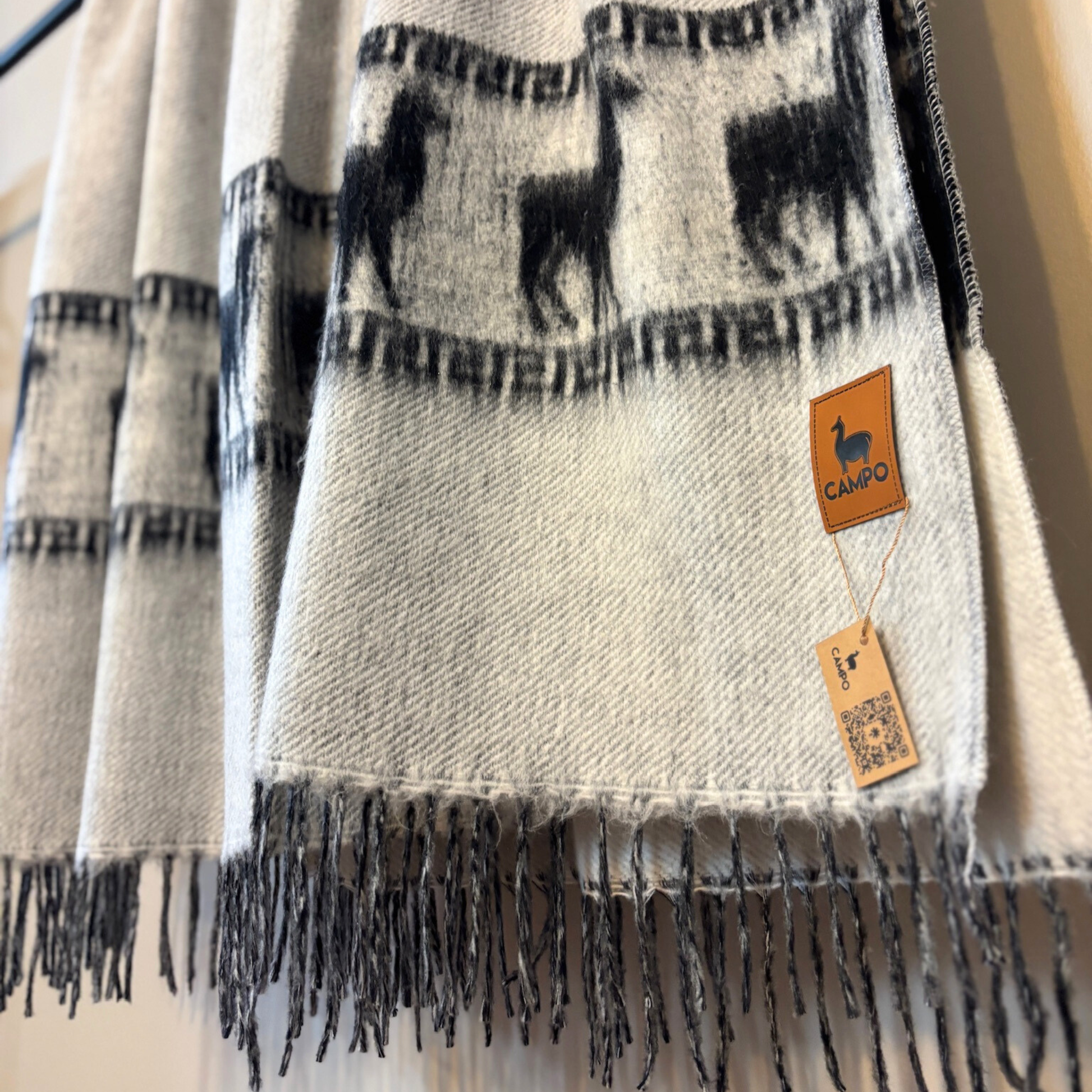 light gray scarf with black patterns and fringes, featuring a brand tag, on a neutral background.