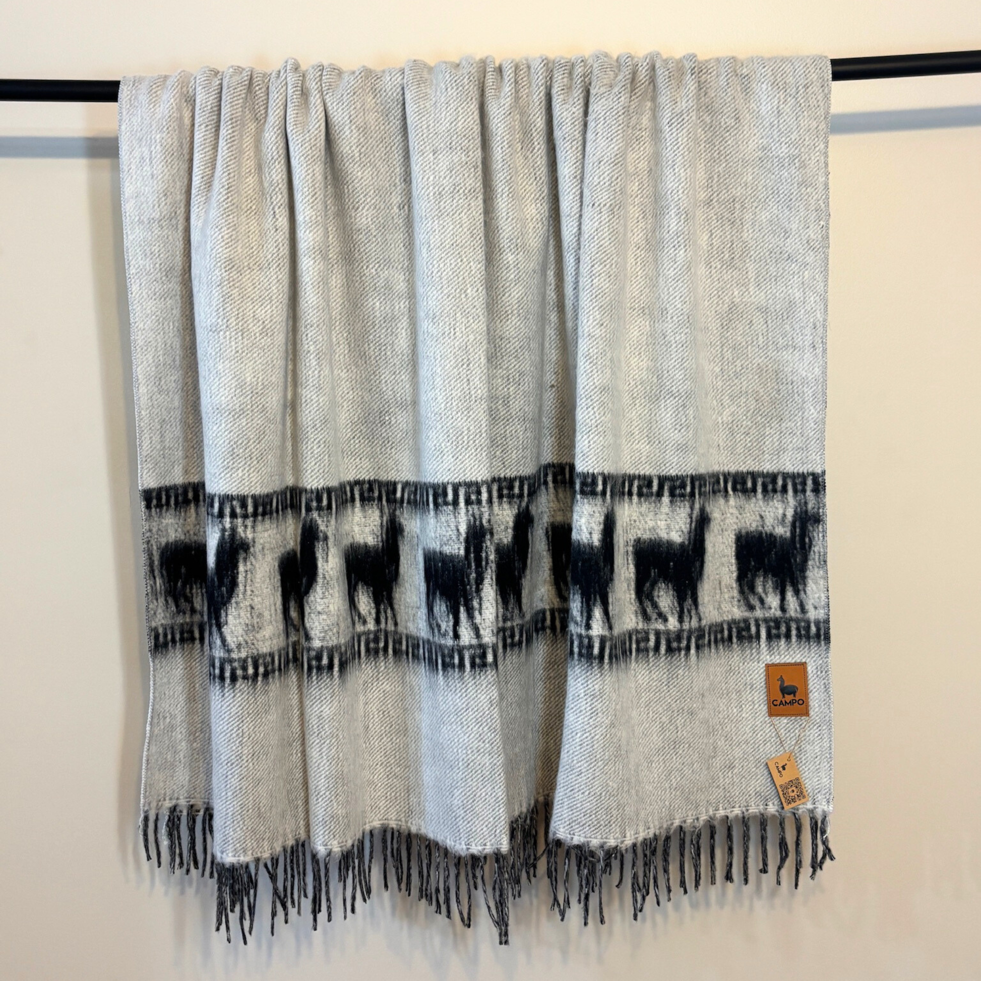 Gray blanket with black alpaca pattern and fringes on a black rack against a white background