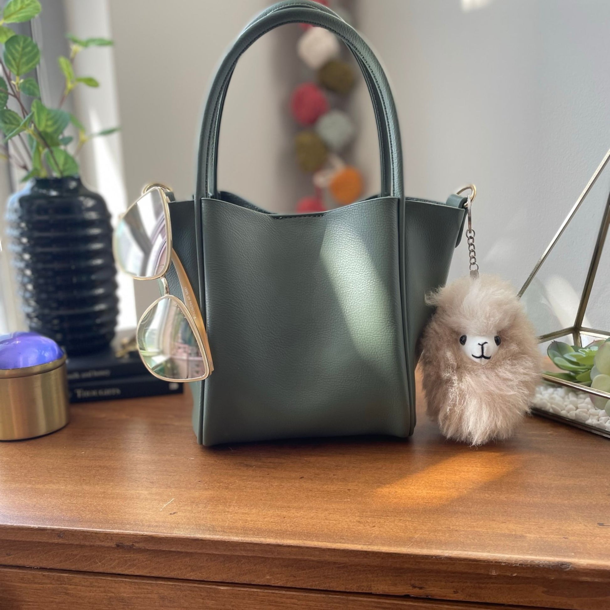 Green handbag on a wooden surface with sunglasses and a fluffy keychain.