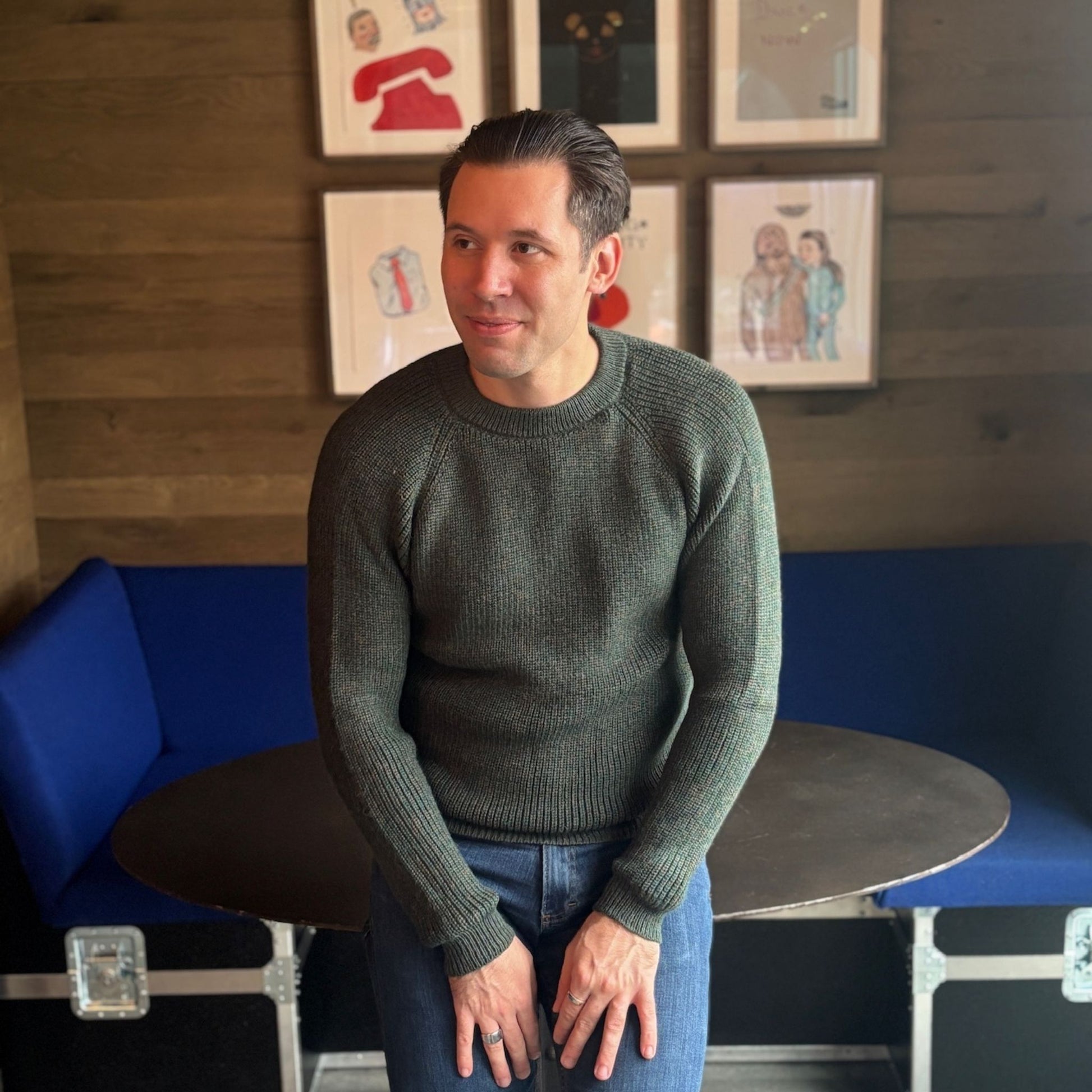 Man in a green sweater sitting on a blue couch with framed pictures on the wall.