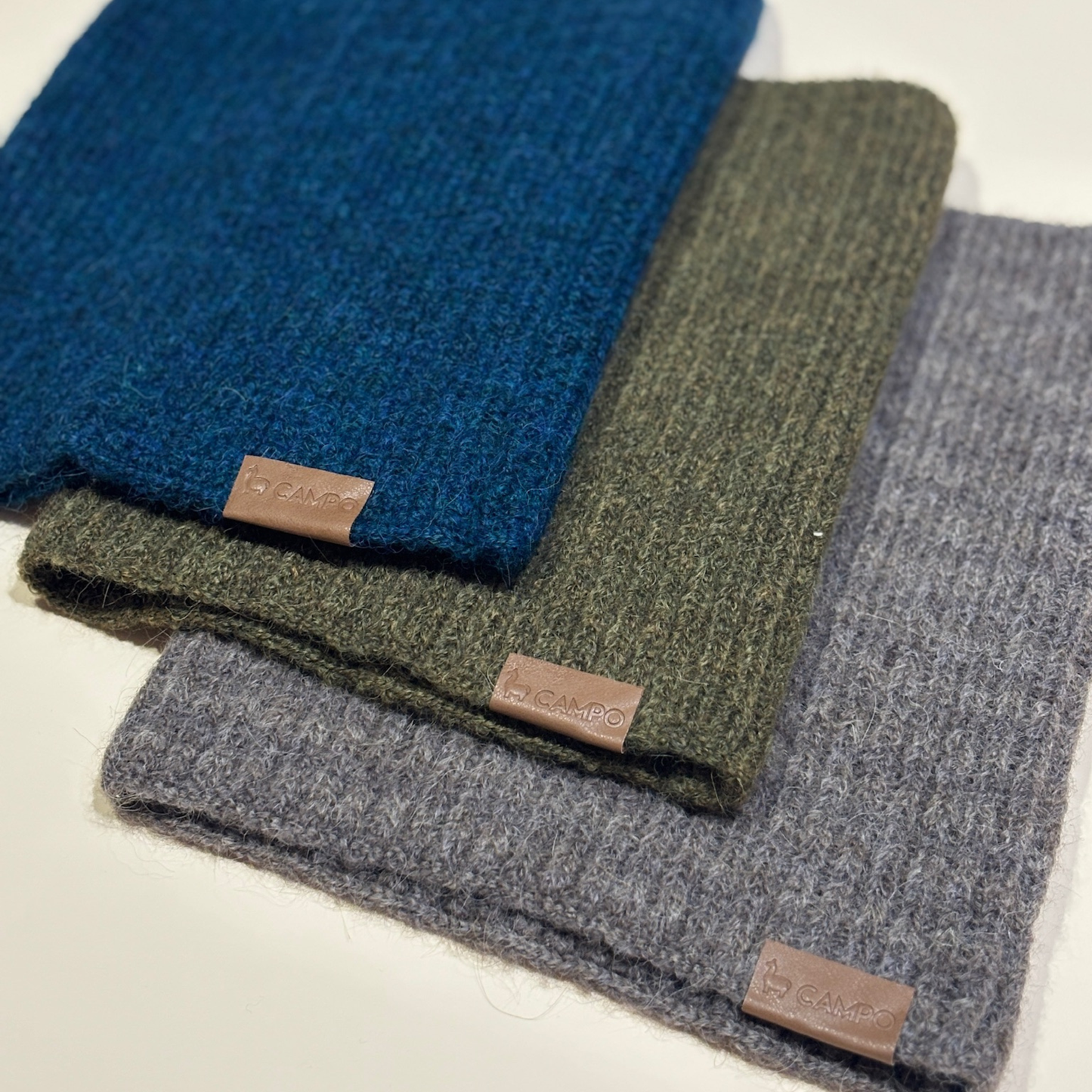 Three folded scarves in blue, green, and gray with visible brand labels on a light background.