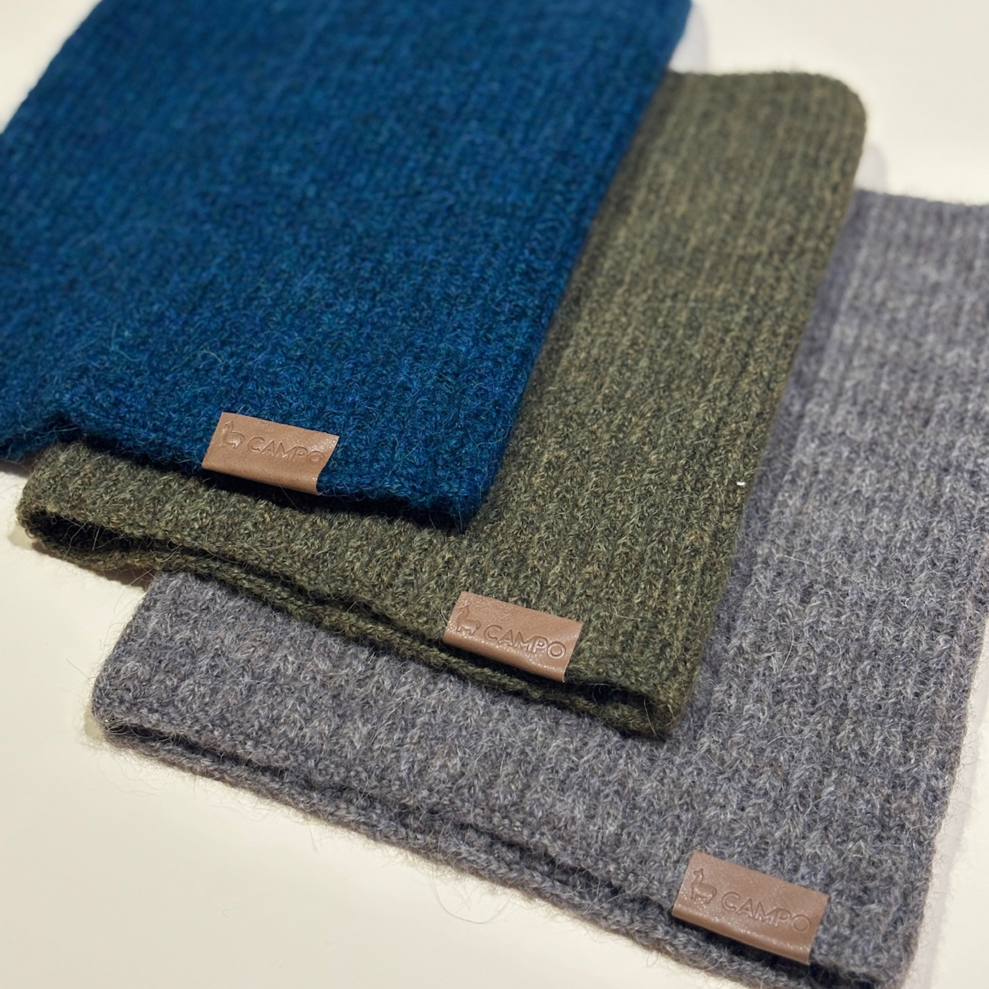 Three folded scarves in blue, green, and gray with visible brand labels on a light background.