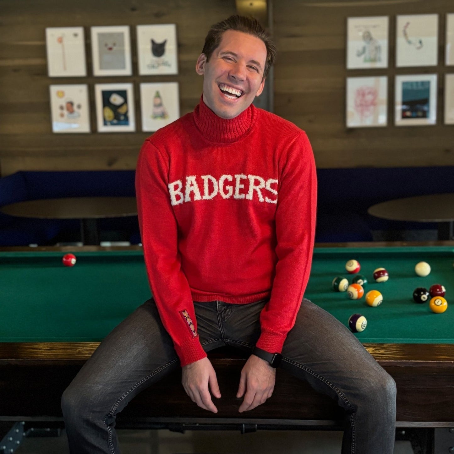 Man wearing a red sweater with 'BADGERS' sitting on a pool table.
