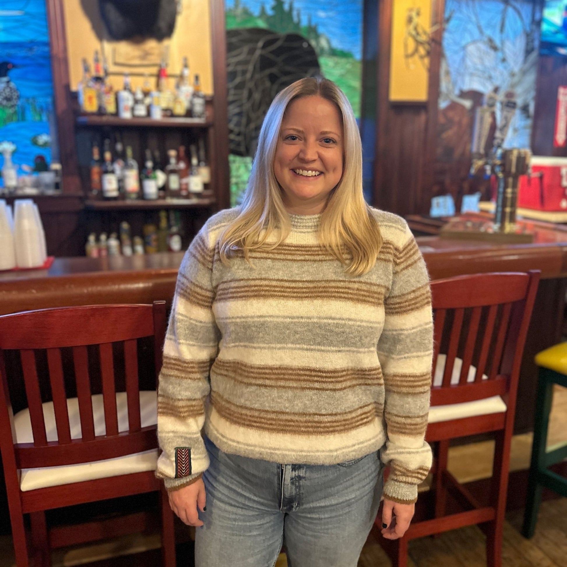 Woman in a striped sweater standing in a bar with wooden furniture and shelves in the background.
