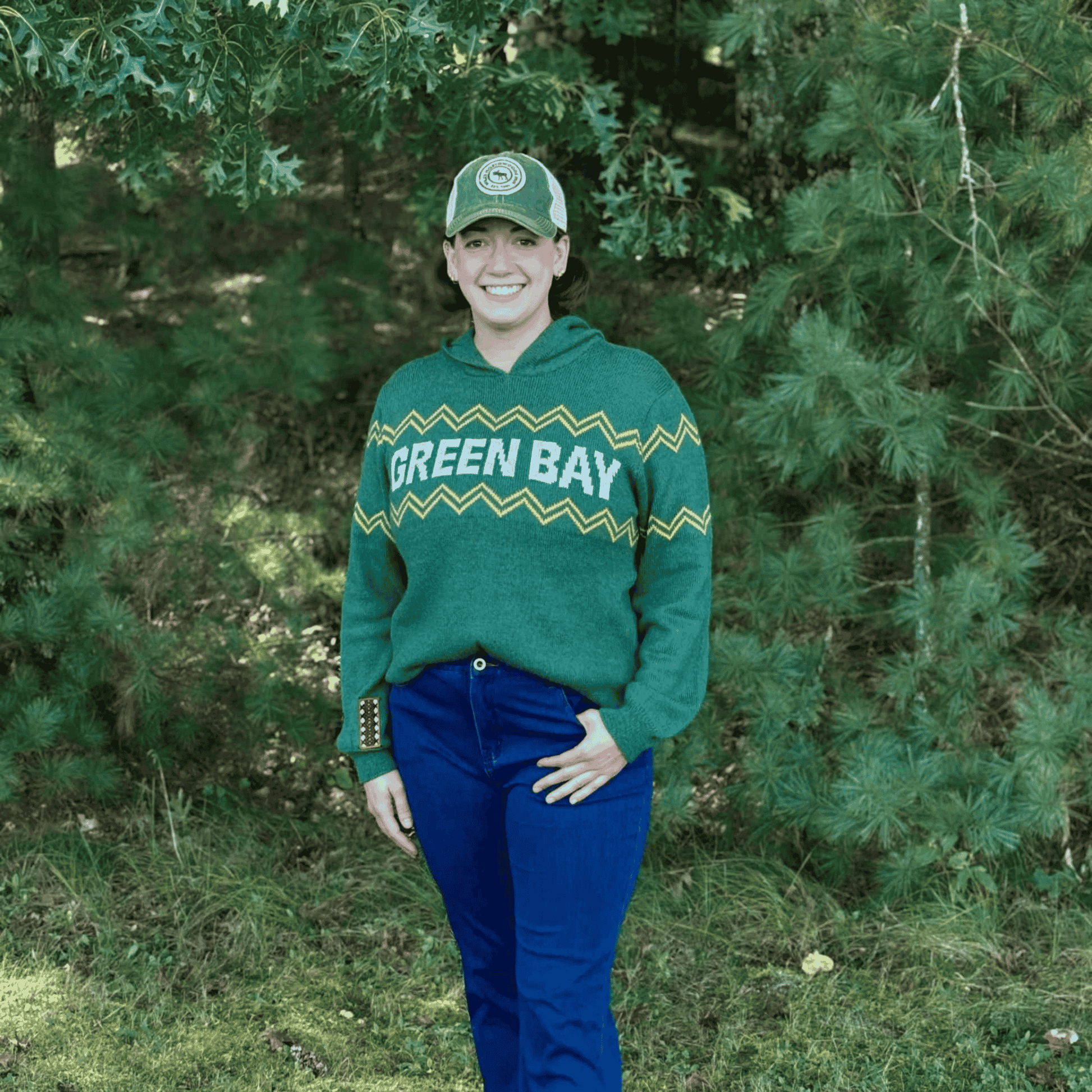 Person wearing a green 'Green Bay' sweater and cap standing in a forest.