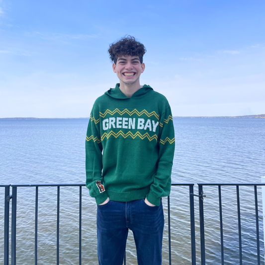 Person wearing a green 'Green Bay' hoodie standing by a waterfront.
