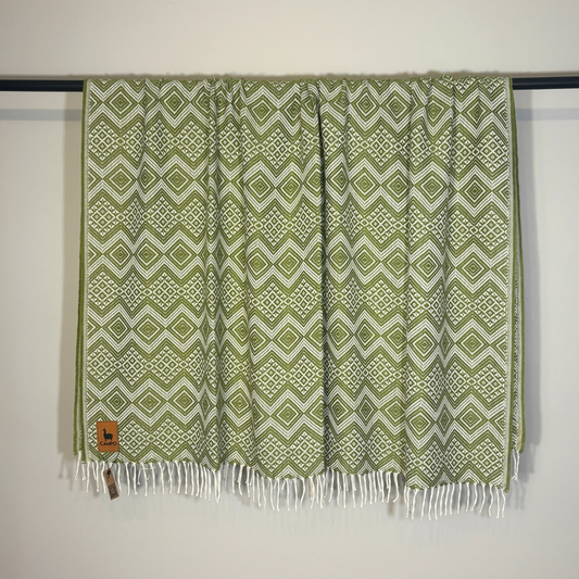 Green patterned throw blanket with white fringe on a black rod against a white wall.