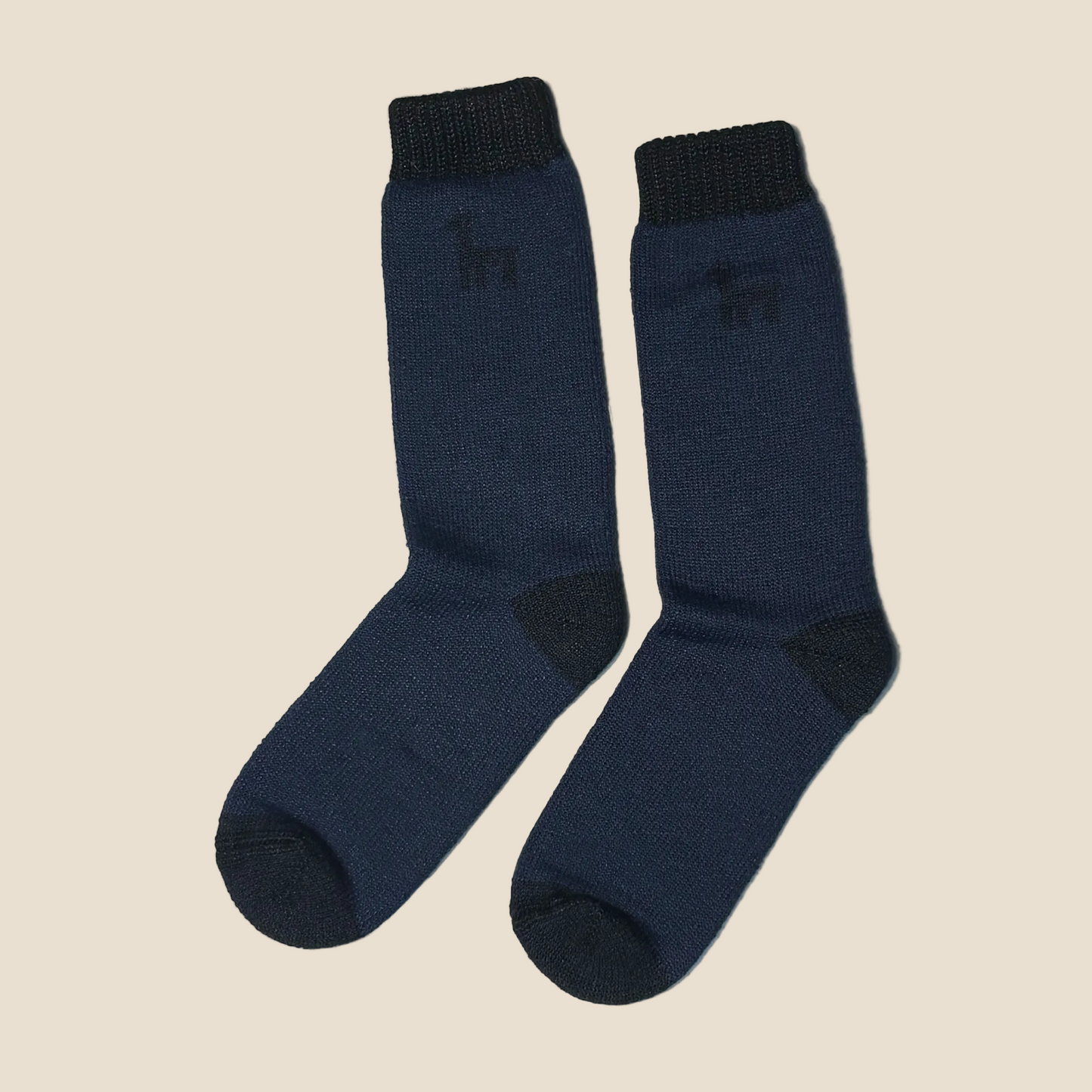 A pair of navy blue crew socks with dark charcoal accents on the heels, toes, and ribbed cuffs, featuring a small embroidered deer logo on the upper leg, set against a solid light beige background.