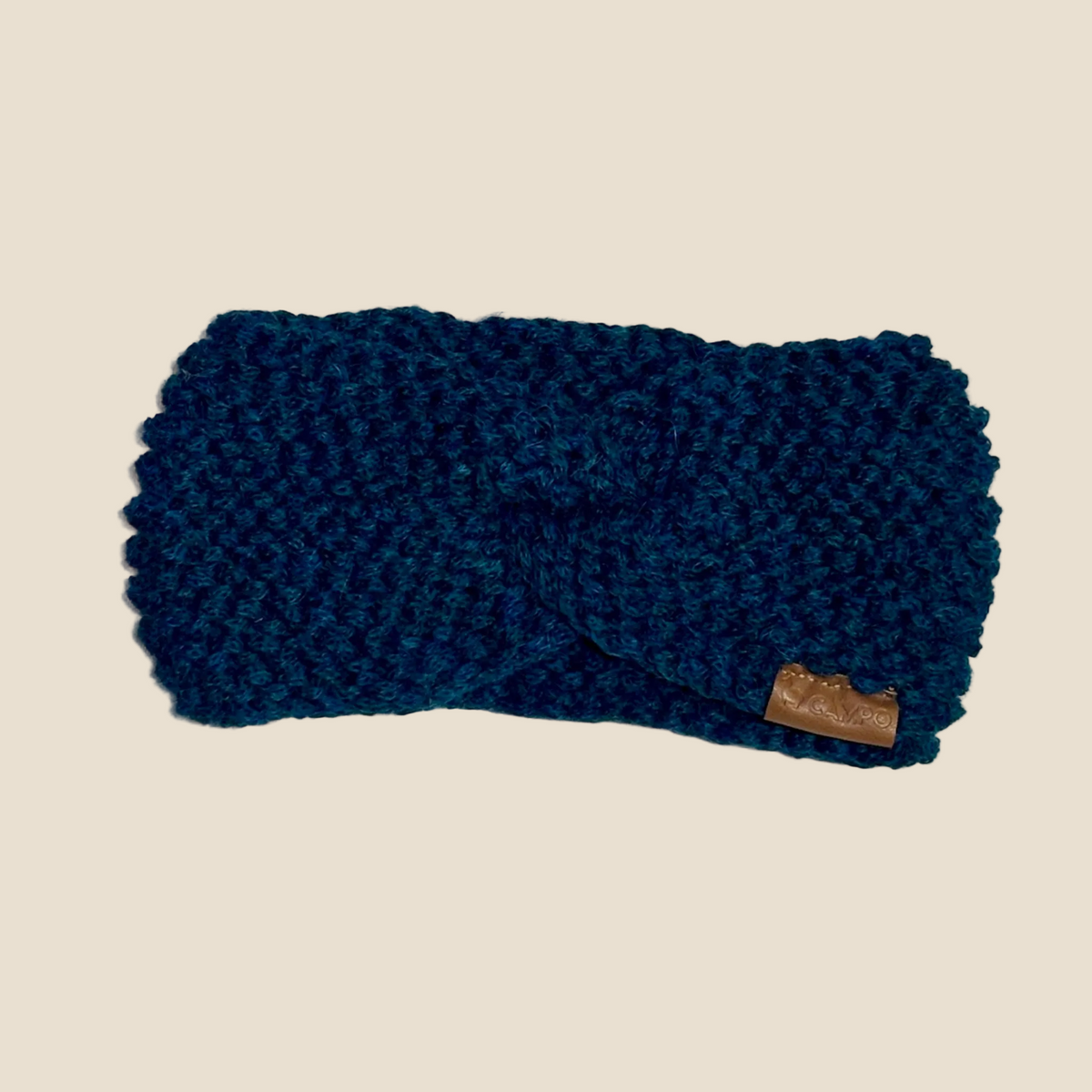 A dark teal blue knitted twist-front ear warmer headband with a small brown leather brand tag, shown on a light textured background.