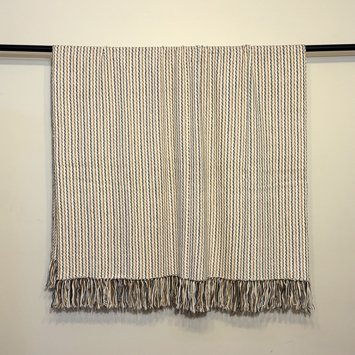 A woven throw blanket with vertical stripes in beige, brown, and gray, hanging from a black rod against a plain off-white wall.