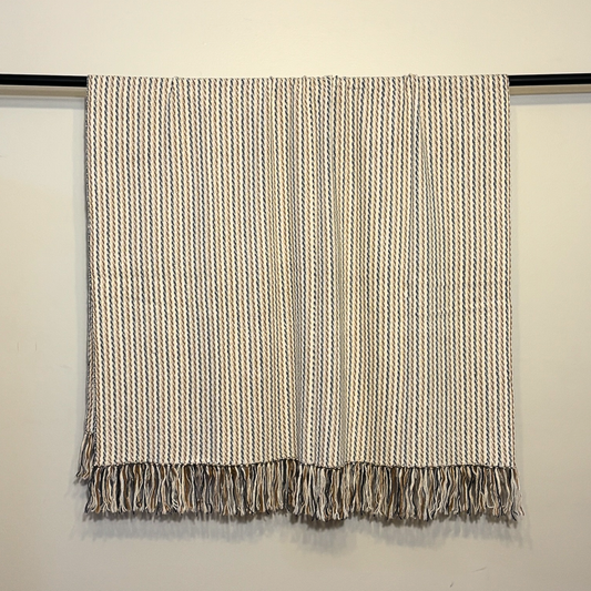 A woven throw blanket with vertical stripes in beige, brown, and gray, hanging from a black rod against a plain off-white wall.