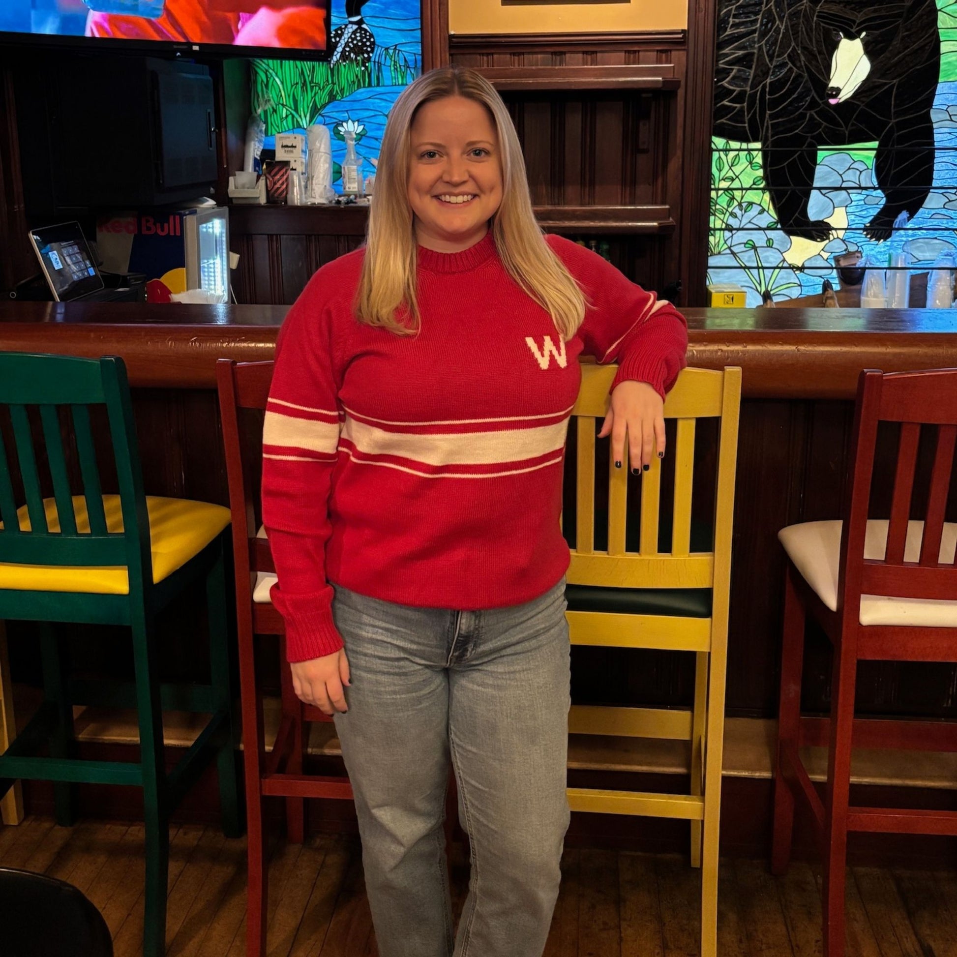 Woman wearing a red sweater with a 'W' logo in a casual setting with chairs and a stained glass window.