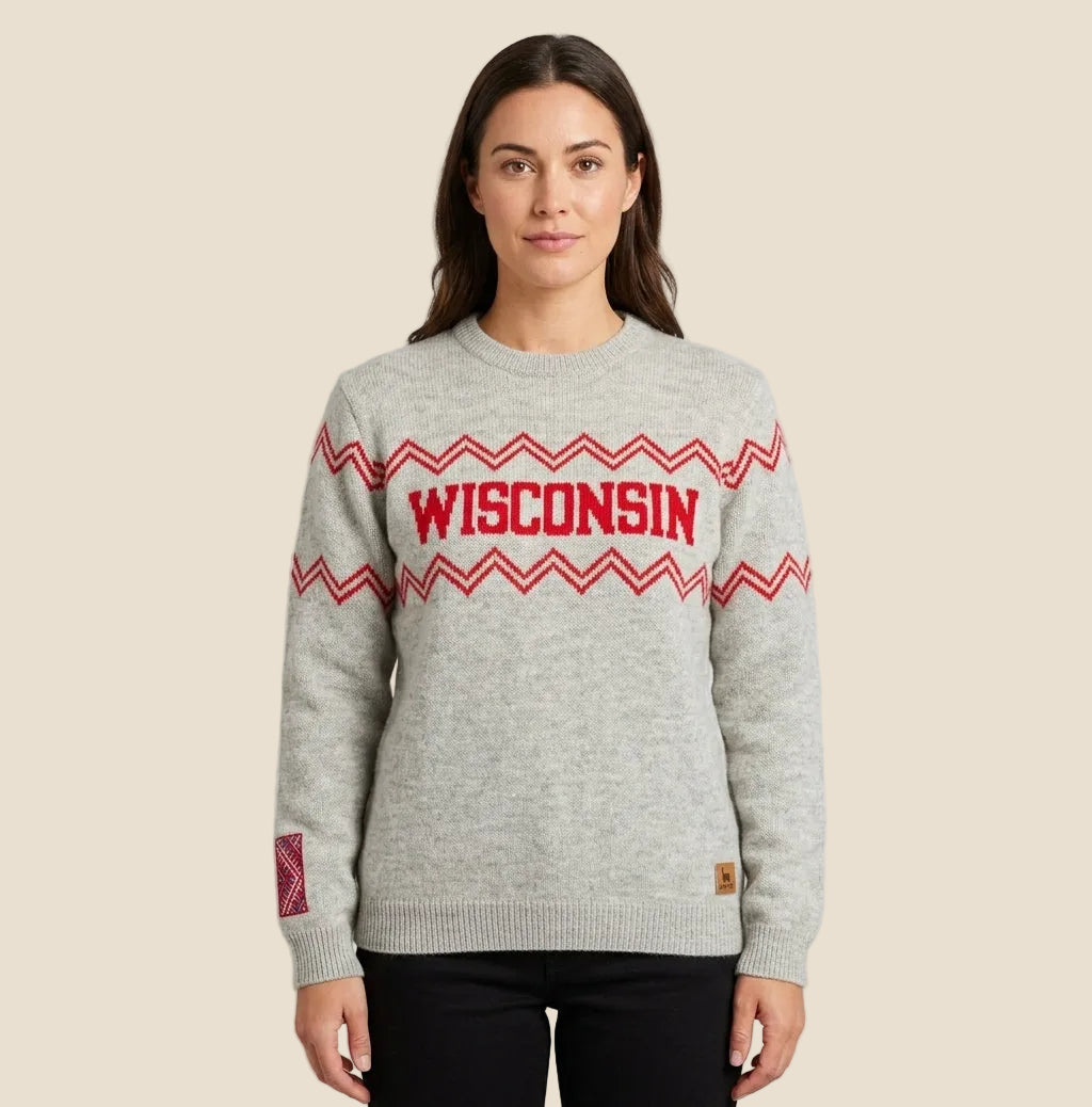 A woman modeling a light gray heather crewneck sweater with red and white zigzag patterns and 'WISCONSIN' printed across the chest in red, paired with black pants against a light gray studio background.