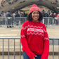 Person wearing a red 'Wisconsin' sweater and pink beanie in an outdoor setting with a bridge and people in the background.