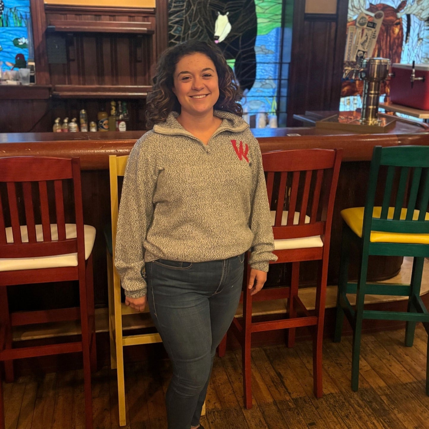 Person wearing a beige sweater with a red logo in a bar setting
