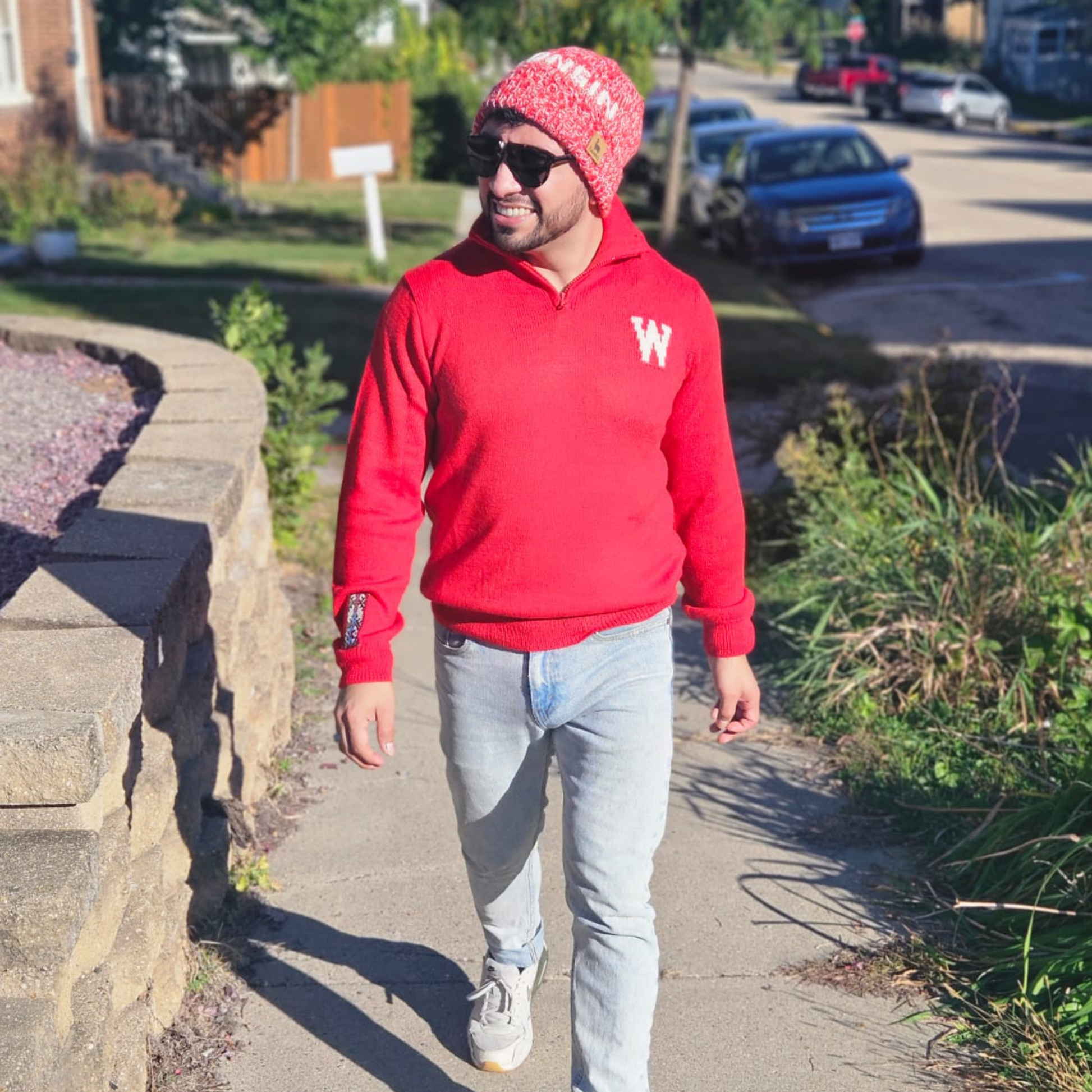 Man in a red hoodie and patterned beanie walking on a sidewalk in a residential area.